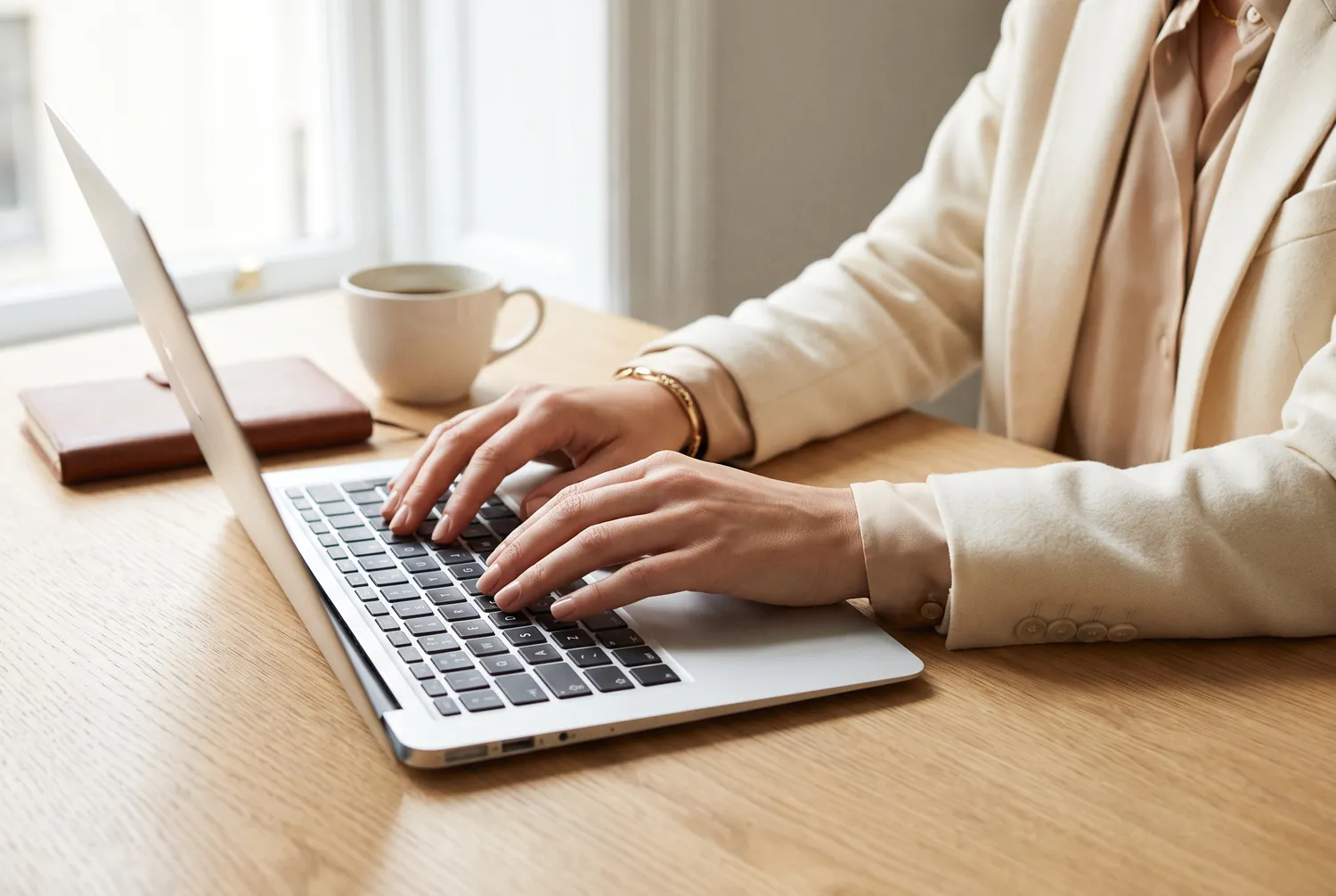 Hände an einem Laptop neben Notizbuch und Kaffeetasse